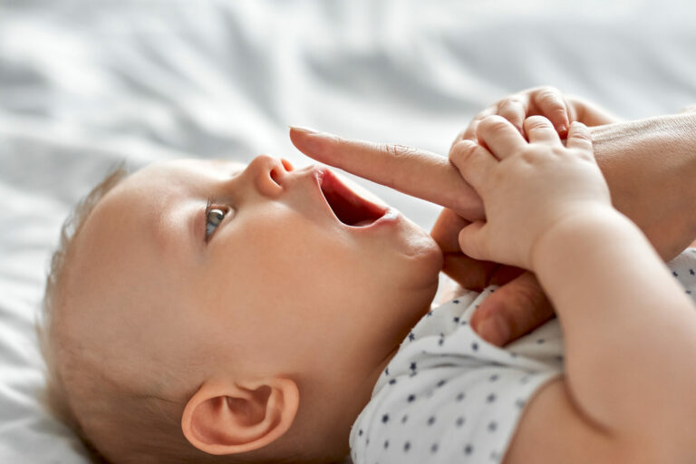 A white baby laying on a sheet, opening its mouth. The baby is grasping the hand of an adult, who has their pointer finger extended over the baby's open mouth.