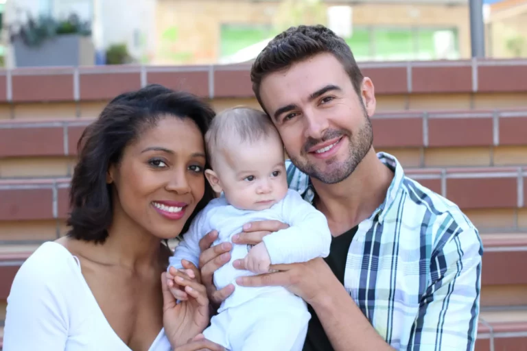 A couple smiling into the camera and holding a baby up in between them.