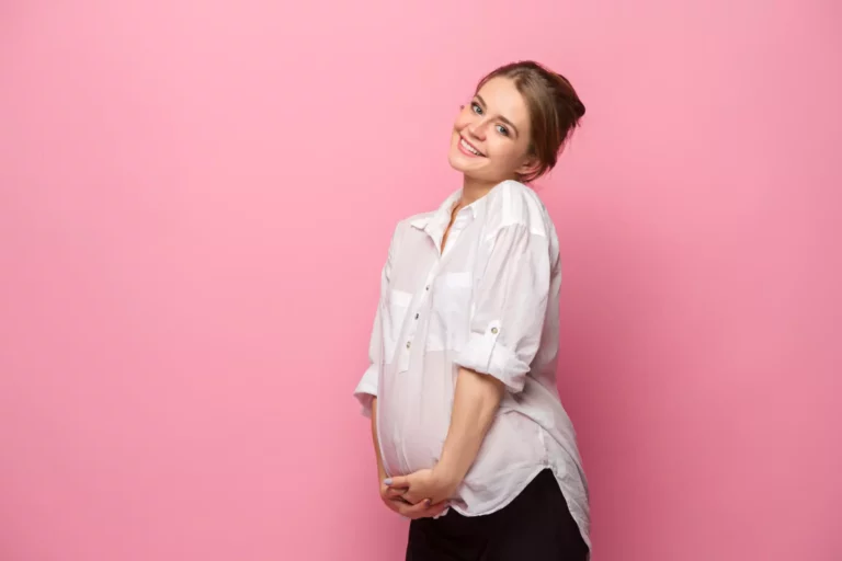 A pregnant woman standing in front of a pink background.