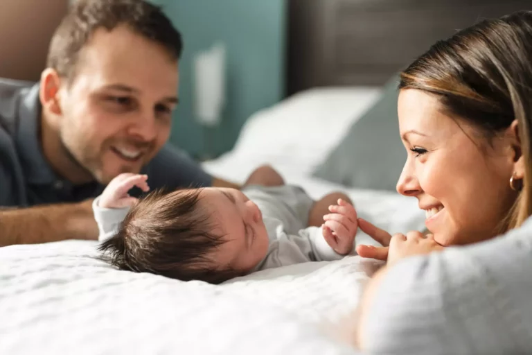 A smiling man and woman are kneeling next to a bed. A baby is laying on the bed in between them.