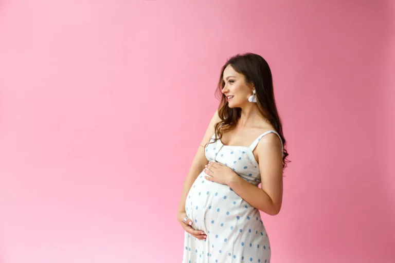 A pregnant woman holding her stomach, standing in front of a pink background.
