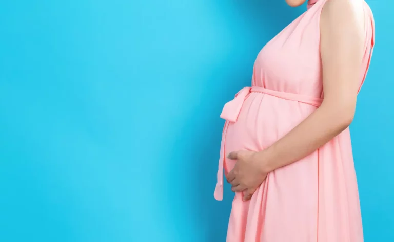 A pregnant woman wearing a pink dress and standing in front of a blue background.