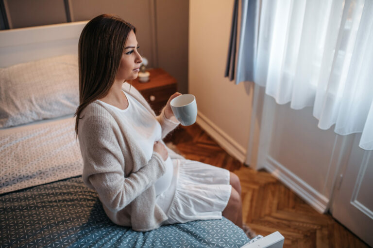 A woman sitting on a bed, holding a coffee mug. She is resting on hand on her pregnant stomach and looking out a window.